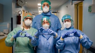 Nurses in Italy dressed in surgical gowns, gloves and protective gear making heart signs to the camera