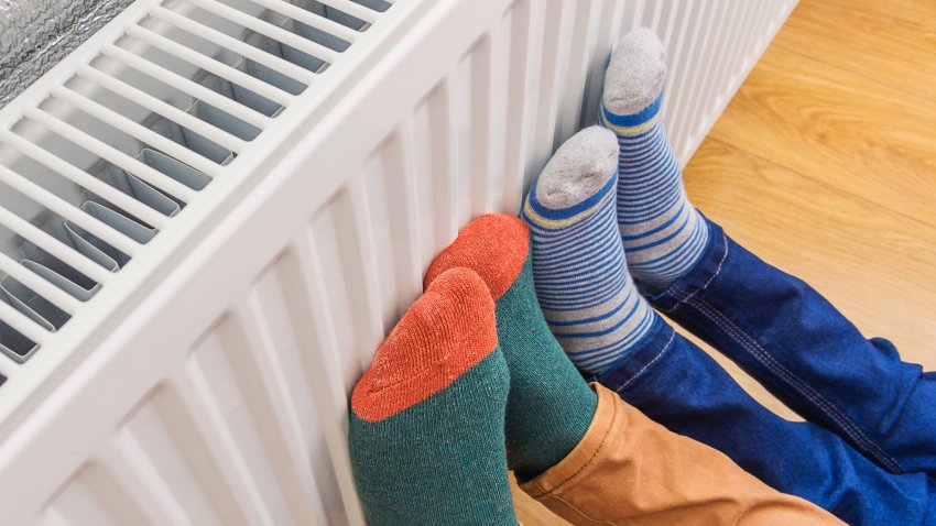 Woman and child wearing colorful pair of woolly socks warming cold feet in front of heating radiator in winter time. Electric or gas heater at home. Part of body, selective focus.