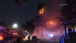 Firefighters are shown in the foreground as a massive church burns in the background, with smoke at the bottom of the frame and flames leaping from a bell tower at the top of the photo.