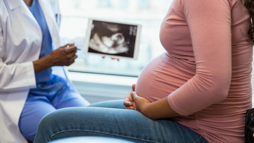 A photo with the focus on the unrecognizable pregnant woman in the foreground as the unrecognizable doctor shows her an ultrasound on a digital tablet in the background.