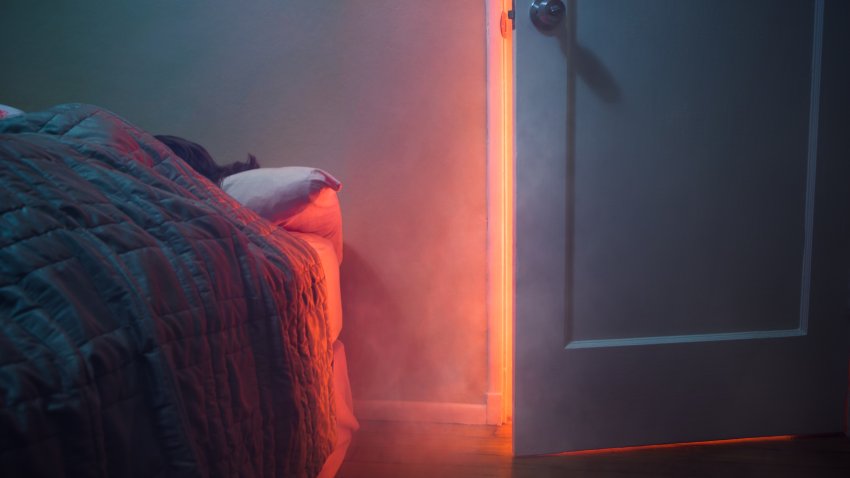 A depiction of a house fire, indoors.  Smoke and light from a fire pour through a cracked room door, a person visible sleeping in their bed.  Horizontal with copy space.