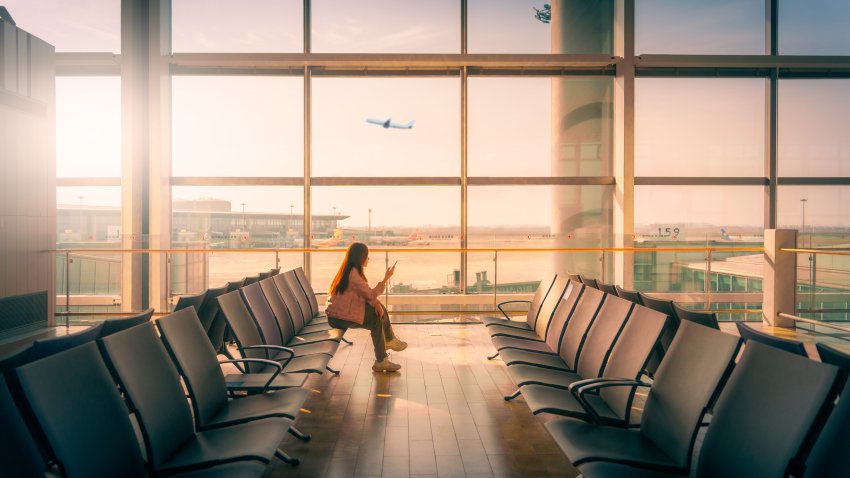 A confident young Asian businesswoman with a suitcase uses her smartphone while waiting for her flight at an airport terminal. The scene reflects modern business travel, professionalism, and mobile connectivity in air transportation.
